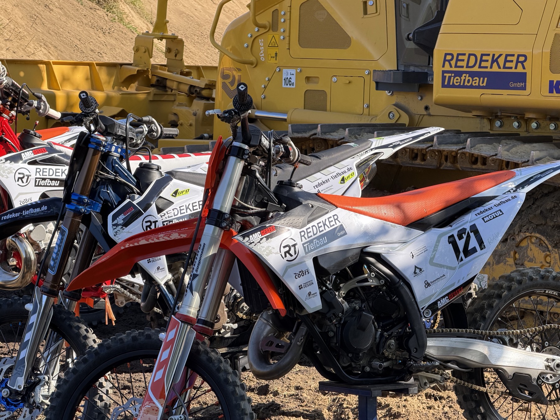 Motocross-Motorrad im Redeker-Tiefbau-Design vor einem Baufahrzeug der Redeker Tiefbau GmbH